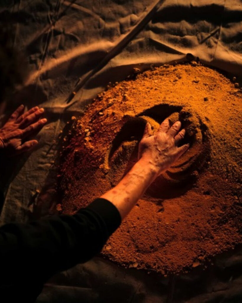 Hands pressing into a mound of red earth, Conjugação Terra at NUMA.SALA